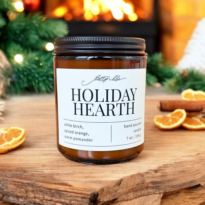Holiday Hearth candle in amber glass jar with black lid, surrounded by dried orange slices and festive greenery