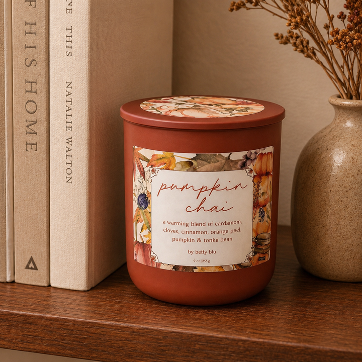 Pumpkin Chai candle by Betty Blu with its floral lid on, styled on a wood shelf beside books and a stoneware vase.