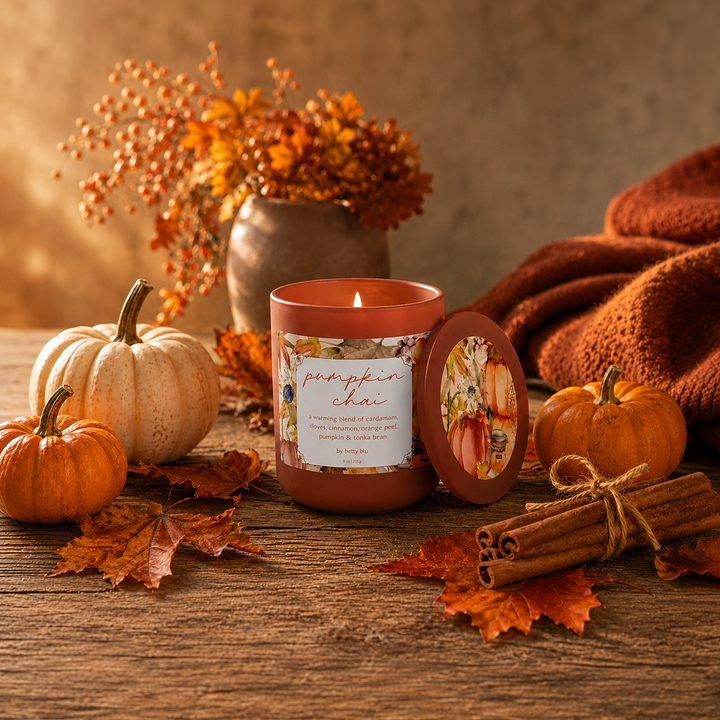 Lit Pumpkin Chai fall candle by Betty Blu on a rustic wood table with mini pumpkins, maple leaves and cinnamon sticks.